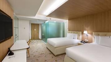 A hotel room with two beds, a desk, a TV, and a bathroom with a glass door.
