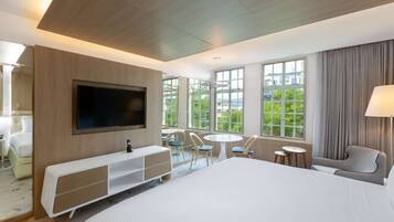 Habitación de hotel moderna con una cama grande, un televisor montado en la pared, un escritorio con una silla, una pequeña mesa redonda con sillas y ventanas amplias que ofrecen vistas al verdor.