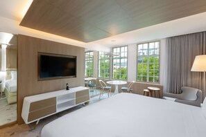 A modern hotel room with a large bed, a TV mounted on the wall, a desk with a chair, a small round table with chairs, and large windows offering a view of greenery. - Gran Hotel Costa Rica, Curio Collection by Hilton (San José)