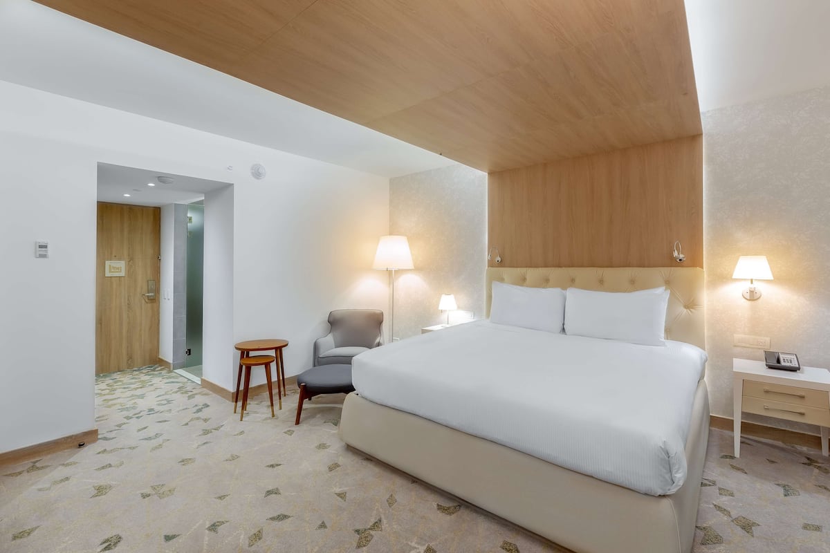 A hotel room with a large bed, two bedside lamps, a chair, a small table, and a wooden door.