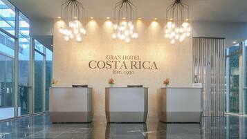 A reception area with a modern design, featuring three reception desks, a chandelier, and a wall with the hotel's name and establishment year.