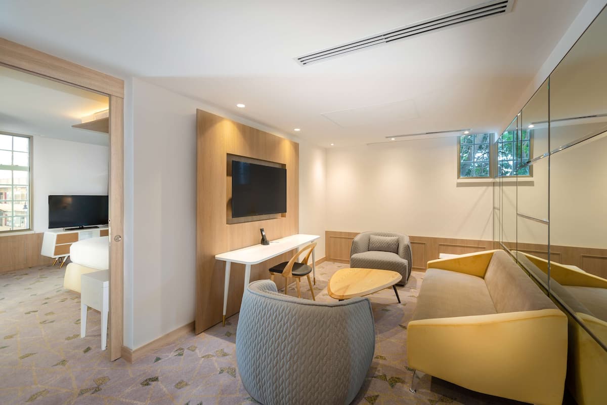 A modern hotel room with a flat-screen TV mounted on the wall, a small desk with a chair, a sofa, and a large window offering a view of the city.