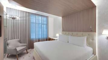 A modern hotel room with a large bed, a seating area with a chair, a desk, and a large window with curtains.