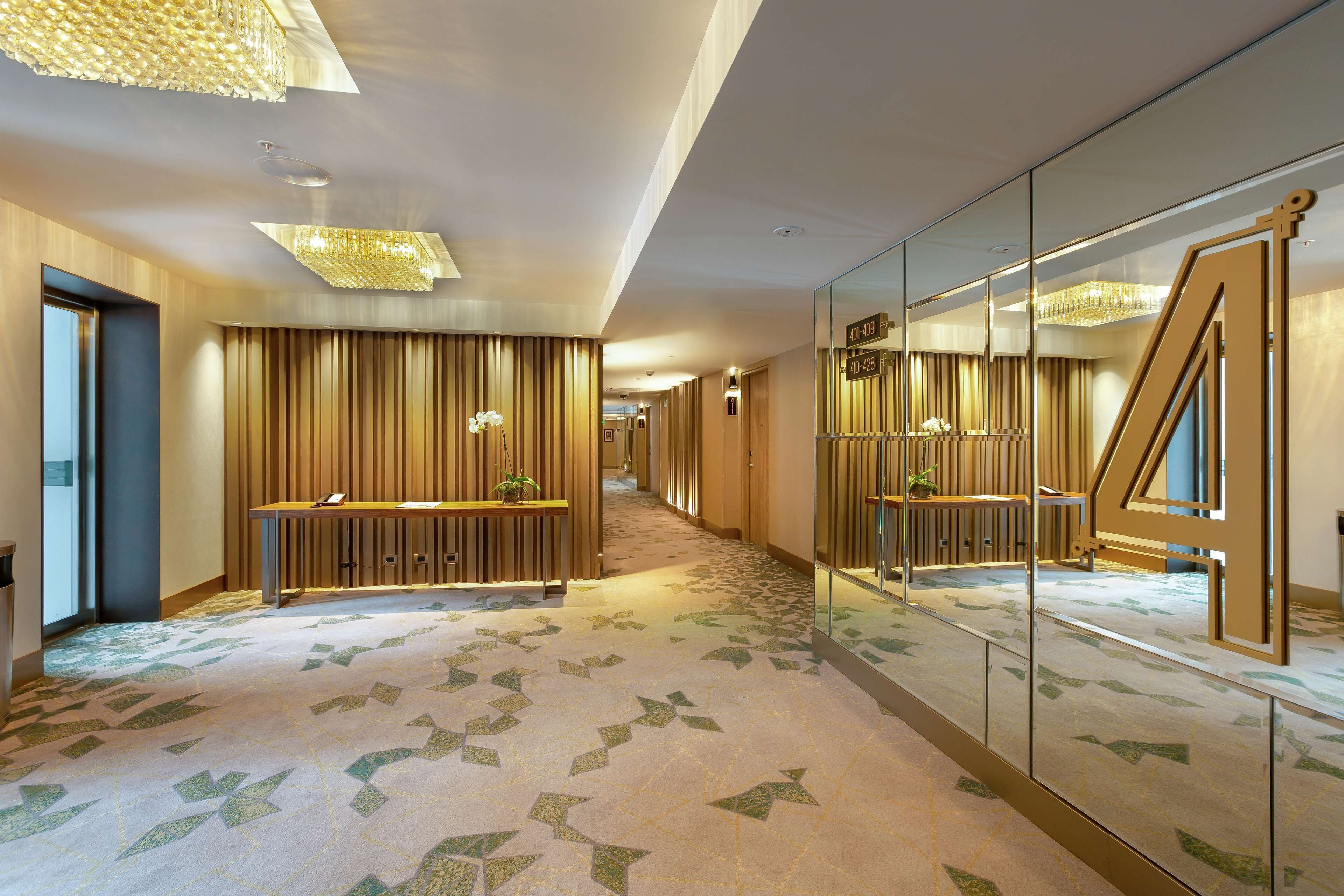 Un pasillo de hotel con alfombra estampada, paneles de madera y una recepción con una planta.