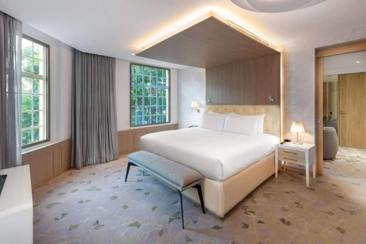 A modern hotel room with a large bed, a bench, a nightstand, and a window with curtains.