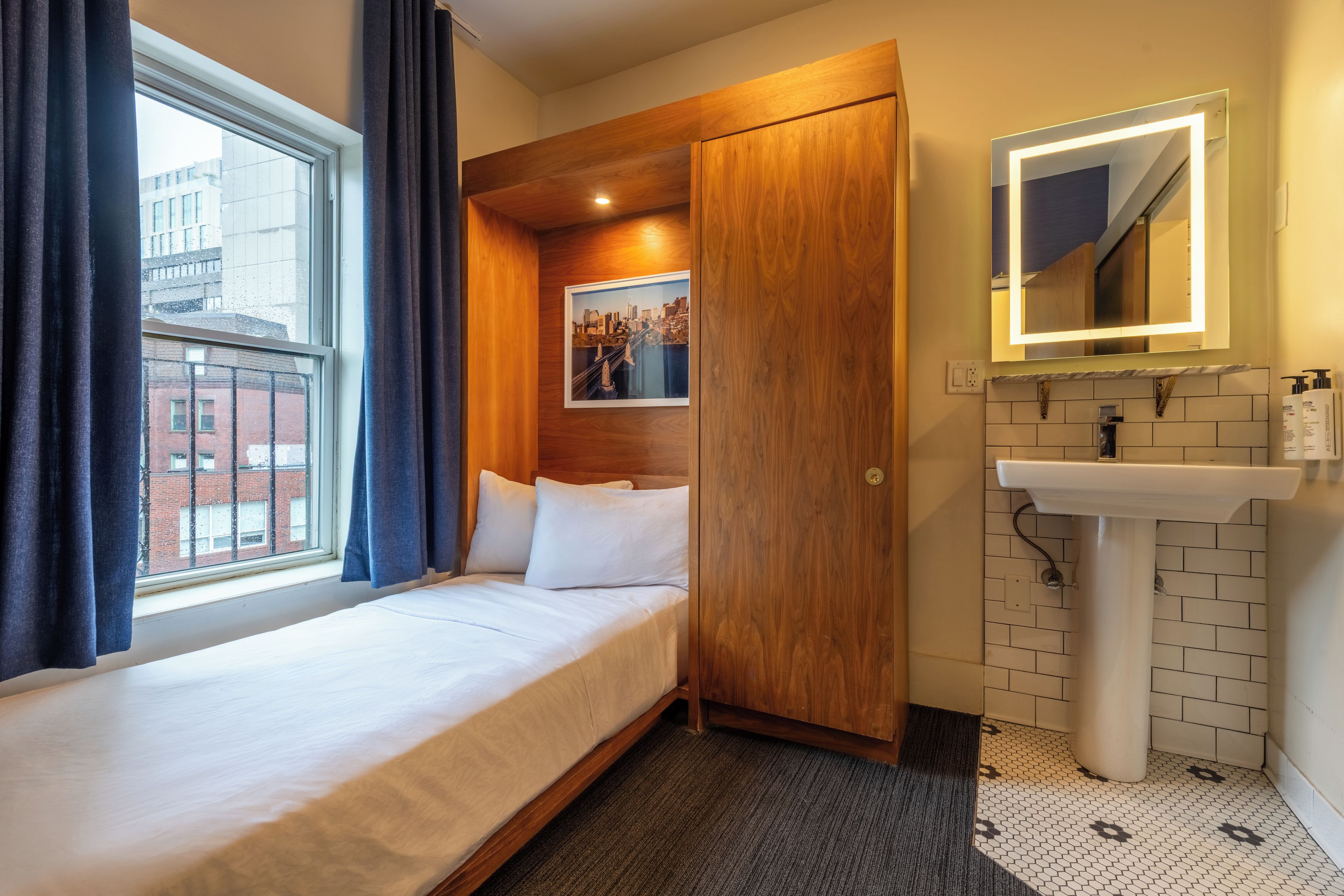 Une chambre d’hôtel comprenant un lit, une armoire en bois, une salle de bain avec un lavabo et un miroir, et une fenêtre avec des rideaux.