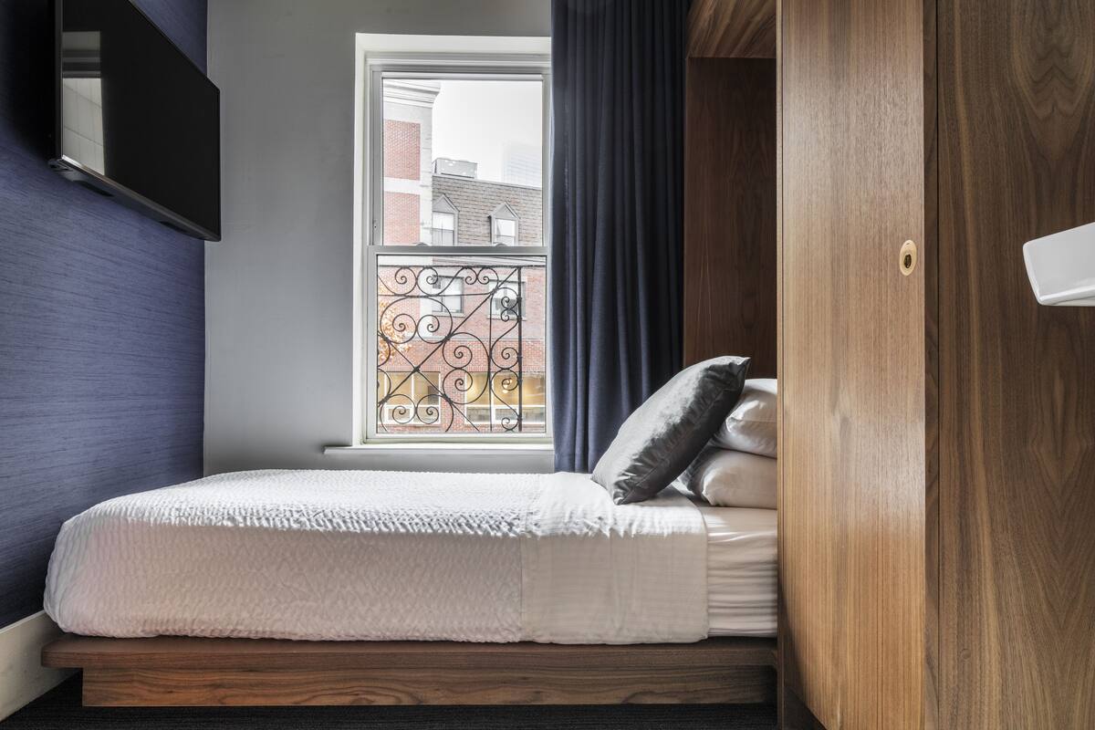 A bedroom with a bed, a window with a view of buildings, and a wooden wardrobe.