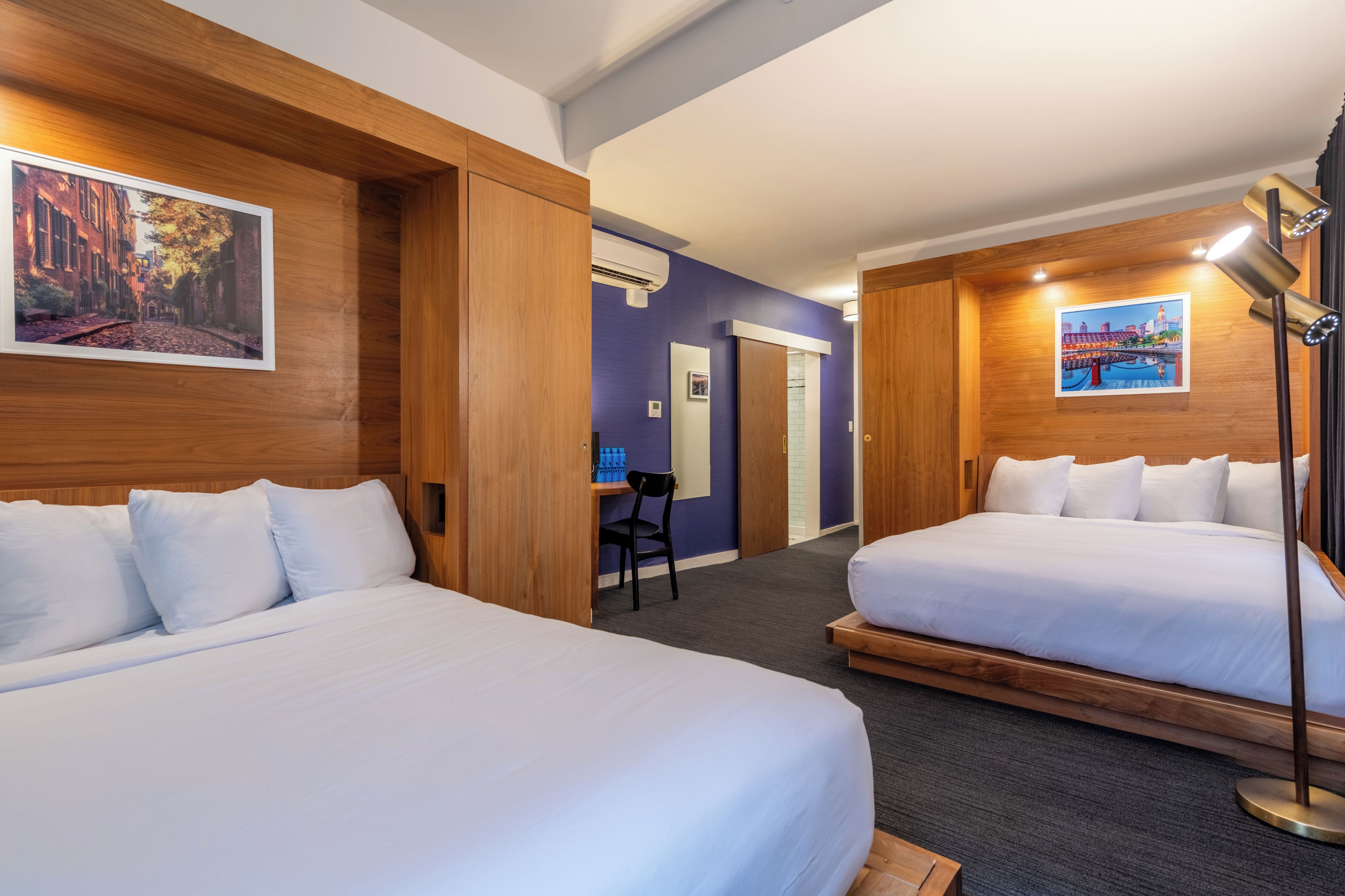 A hotel room with two beds, a desk, and a chair. There are wall-mounted air conditioning units and a lamp.
