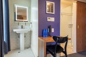Un baño con un lavabo blanco, un espejo, un escritorio de madera, una silla negra y una zona de ducha.