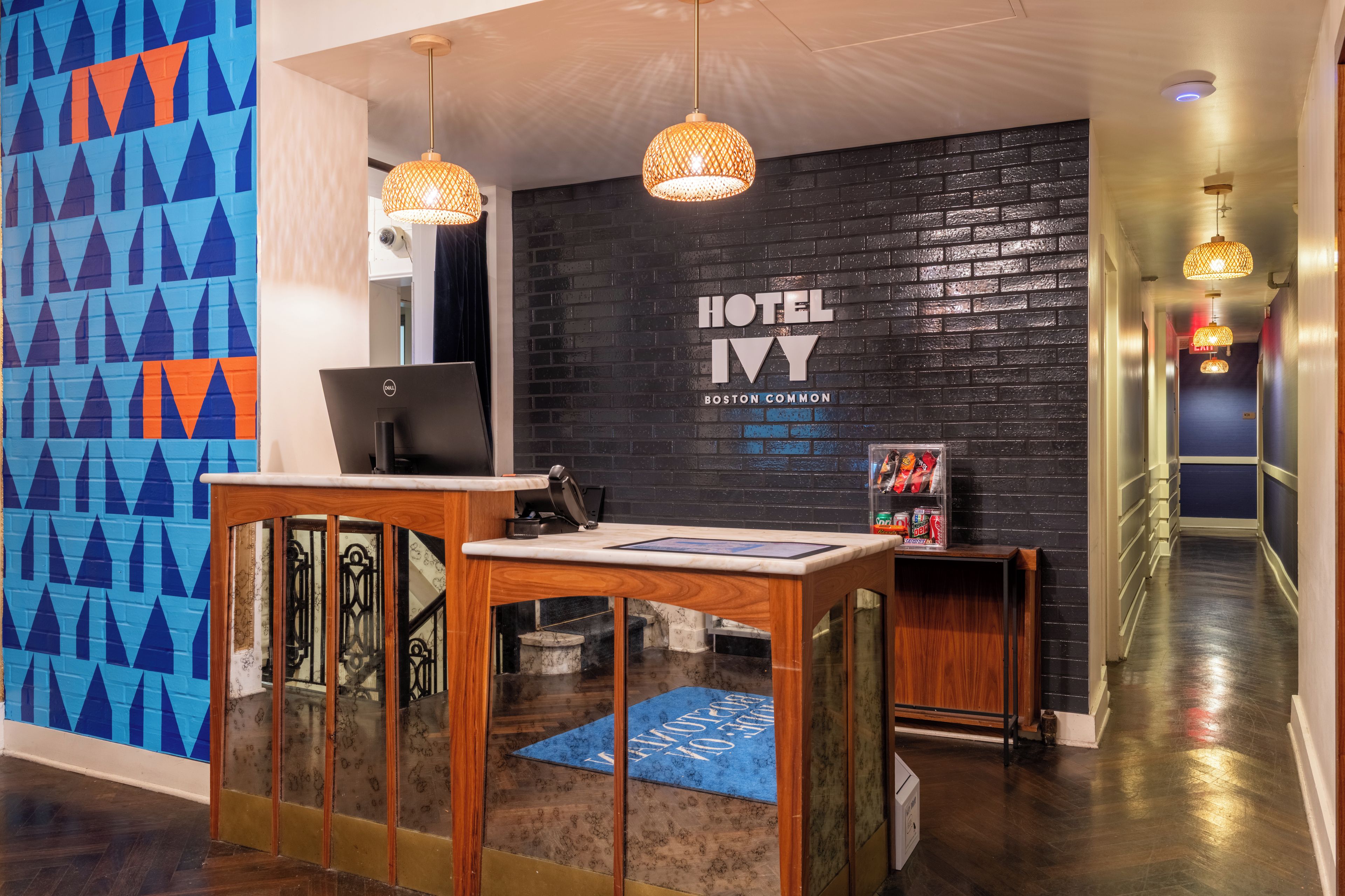 A hotel reception desk with a computer, a wall-mounted sign reading 'HOTEL IVY BOSTON COMMON', and a hallway with a blue carpet leading to a staircase.