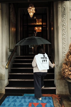 Una persona bajando una escalera con un paraguas negro, vistiendo un chaleco blanco con la inscripción 'HOTEL IVY BOSTON COMMON'.