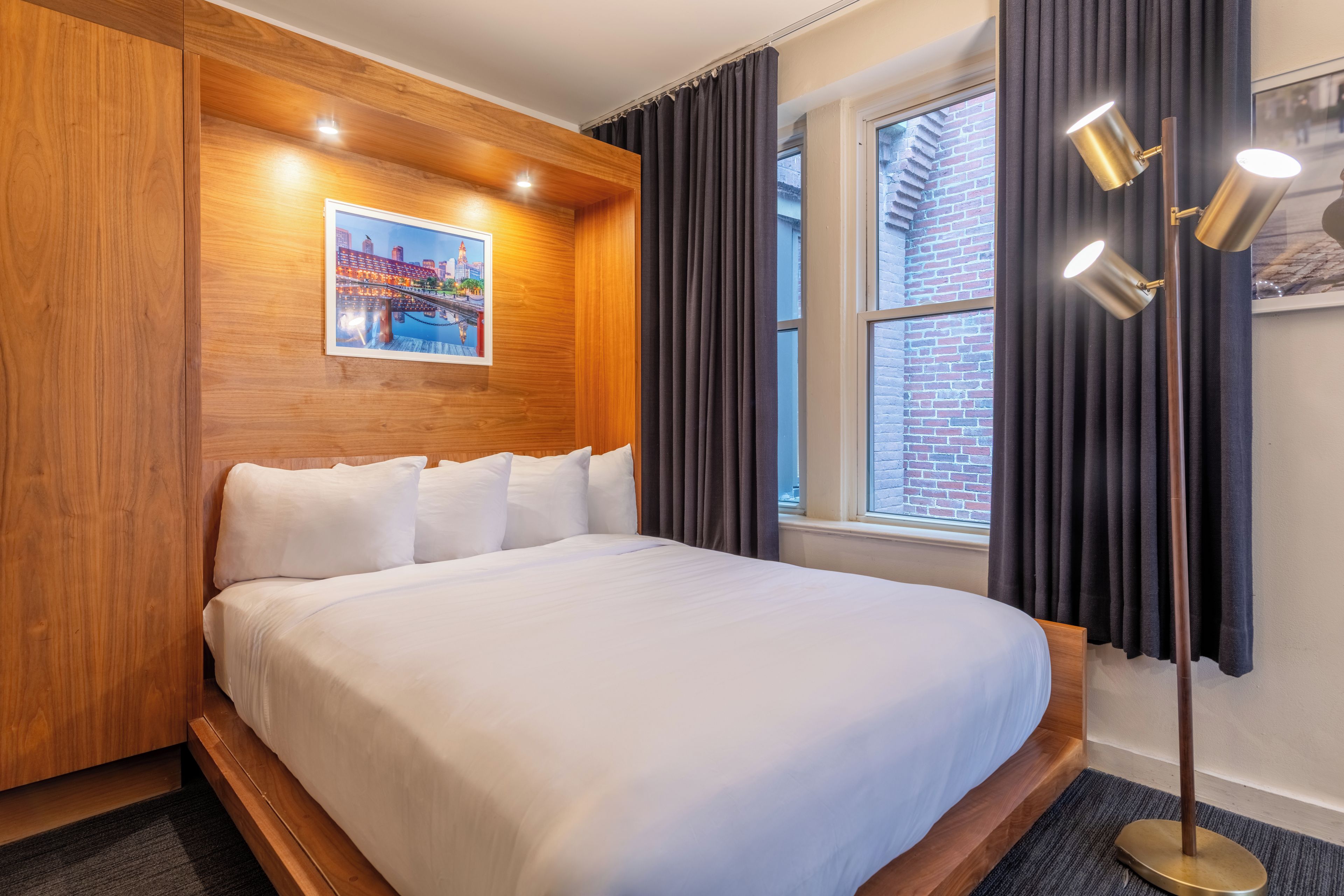 A hotel room with a large bed, wooden headboard, a poster on the wall, and a window with curtains.