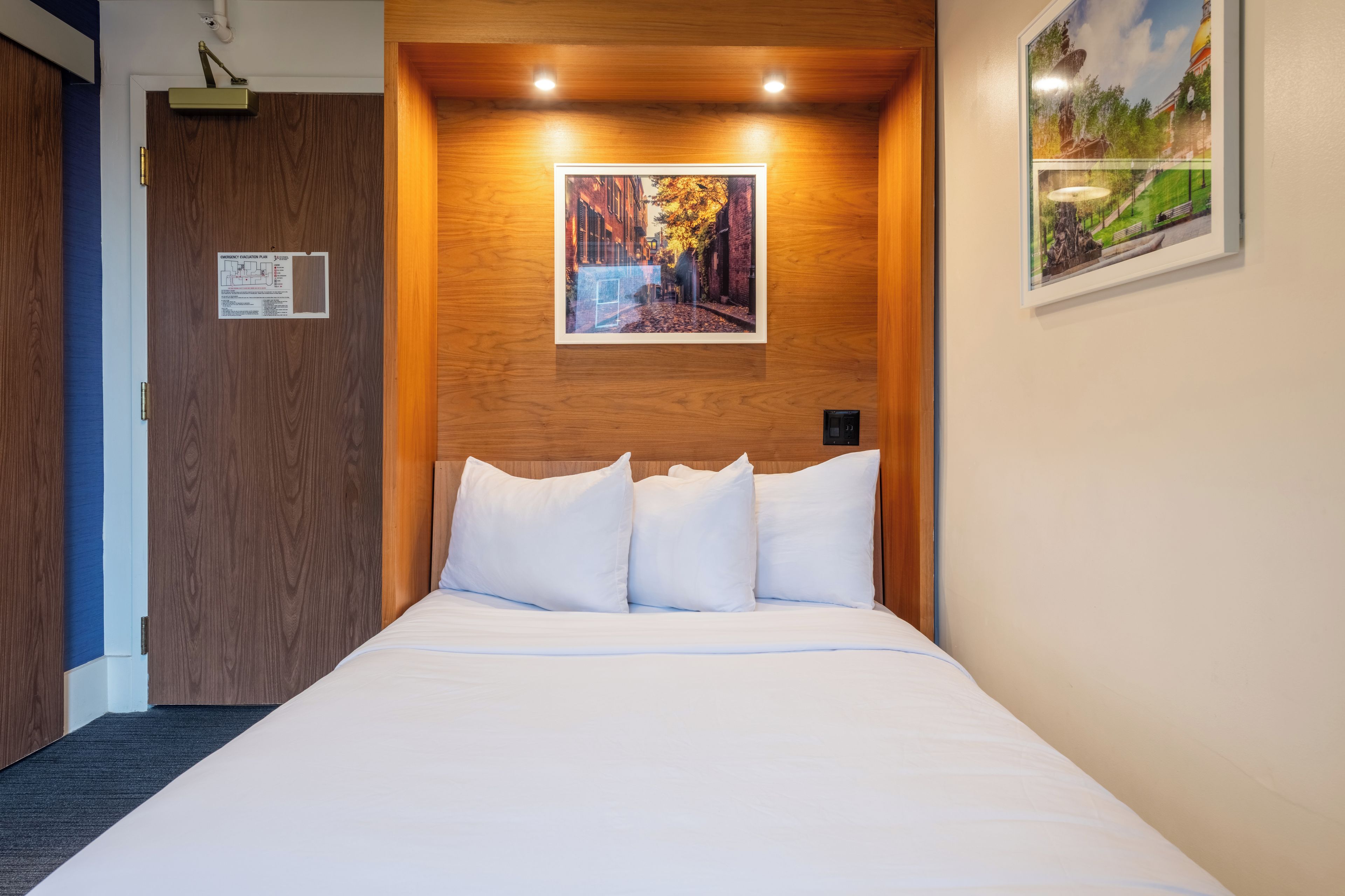 Een hotelkamer met een bed, een houten hoofdeinde, een lijst met een foto aan de muur en een deur met een noodverpakket.
