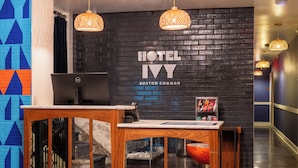 A hotel reception desk with a computer, a wall-mounted sign reading 'HOTEL IV BOSTON COMMON', and a staircase leading to an upper floor.