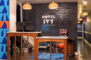 Un comptoir d'accueil d'hôtel avec un ordinateur, une enseigne fixée au mur indiquant « HÔTEL IV BOSTON COMMON », et un escalier menant à l'étage supérieur.