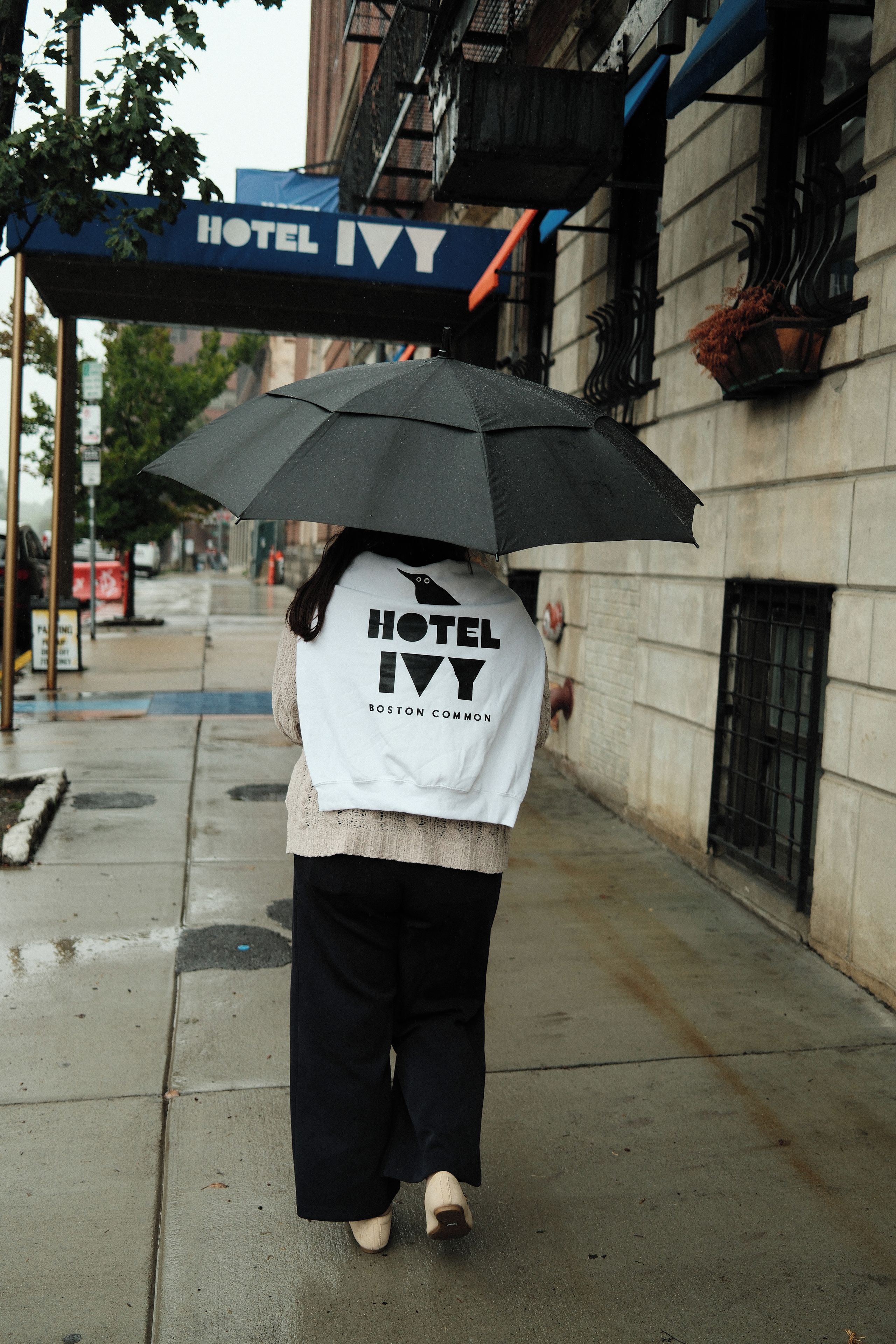 A person walking on a sidewalk with an umbrella, wearing a white jacket with 'HOTEL IVY BOSTON COMMON' text.
