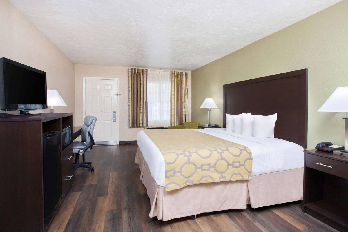 Une chambre d’hôtel avec un grand lit, un bureau avec une télévision, une chaise, une lampe de bureau, une table de chevet avec une lampe et une porte.