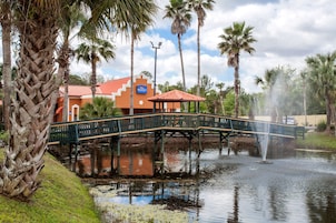 Un puente sobre un estanque con una fuente, rodeado de palmeras y un edificio de techo rojo.