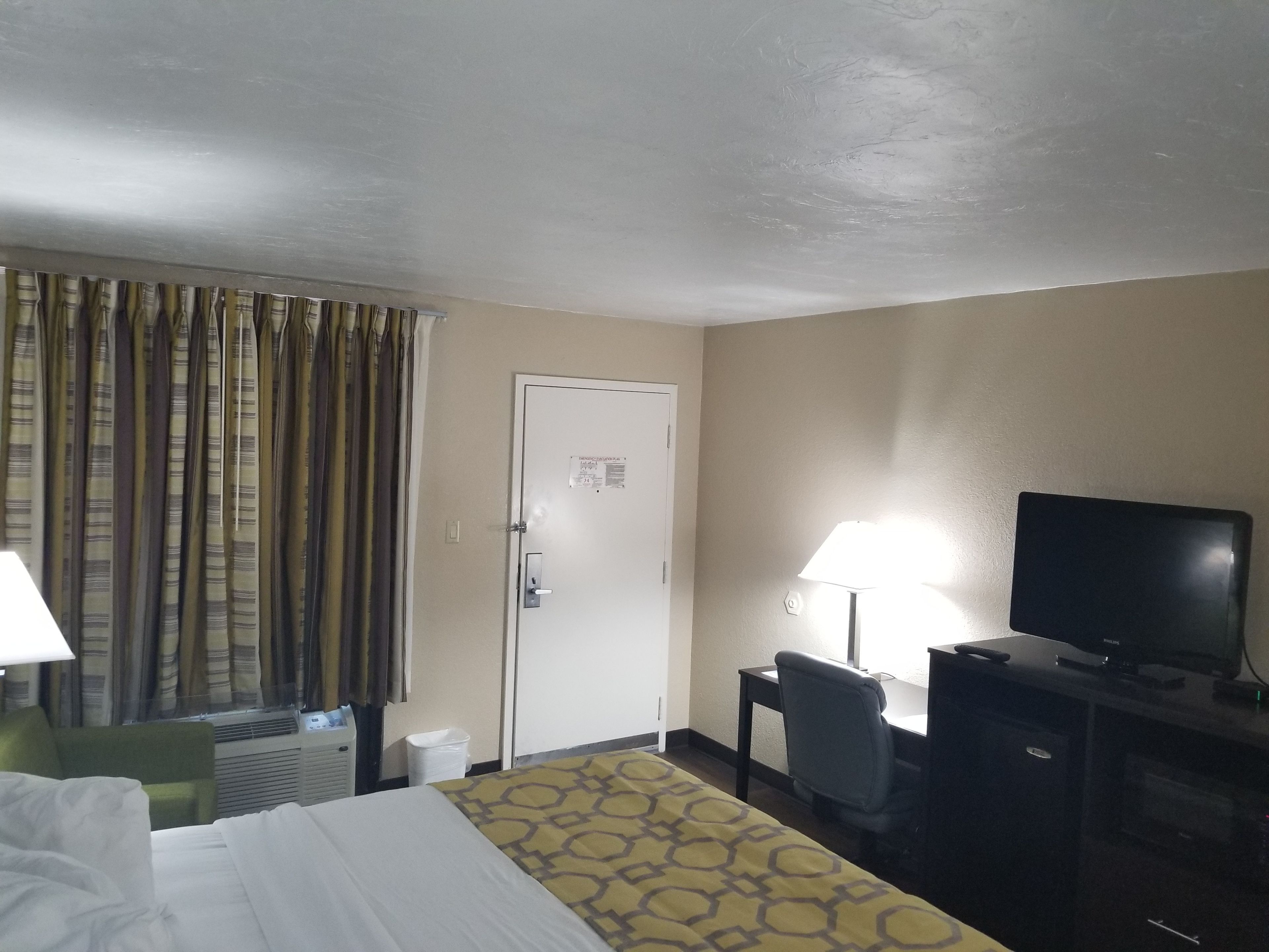 A hotel room with a bed, a desk with a chair, a television, a lamp, and a door.