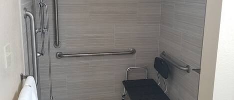Une salle de bain équipée d’une douche, de barres d’appui, d’un banc accessible aux personnes en fauteuil roulant et d’un porte-serviettes.