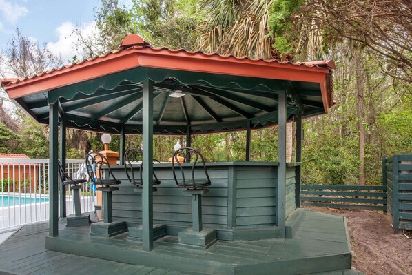 A gazebo with a red roof and green walls, surrounded by trees and a pool.