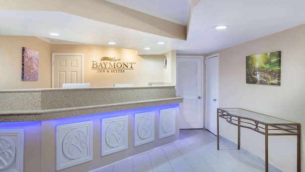 A reception area with a Baymont Inn & Suites sign, a reception desk, a glass-top table, and a door. - Baymont by Wyndham Lake City I-75 (Lake City)
