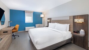 A hotel room with a large bed, a desk, an office chair, a bedside table with a lamp, and a wall-mounted clock.