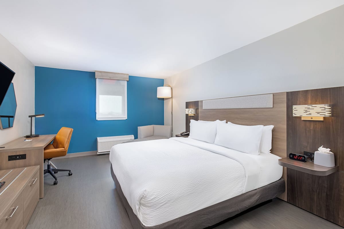A hotel room with a large bed, a desk, an office chair, a bedside table with a lamp, and a wall-mounted clock.