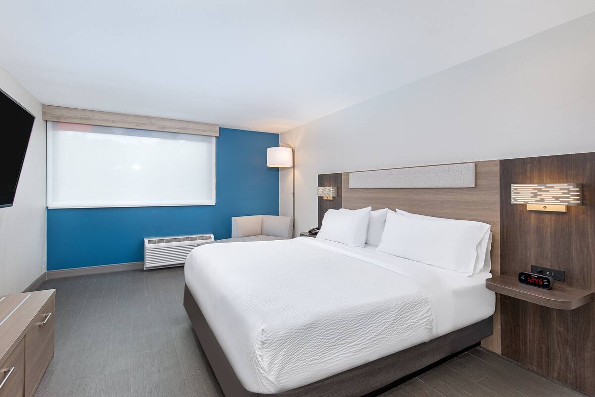 A modern hotel room with a large bed, a bedside table, a nightstand, a lamp, and a wall-mounted clock.