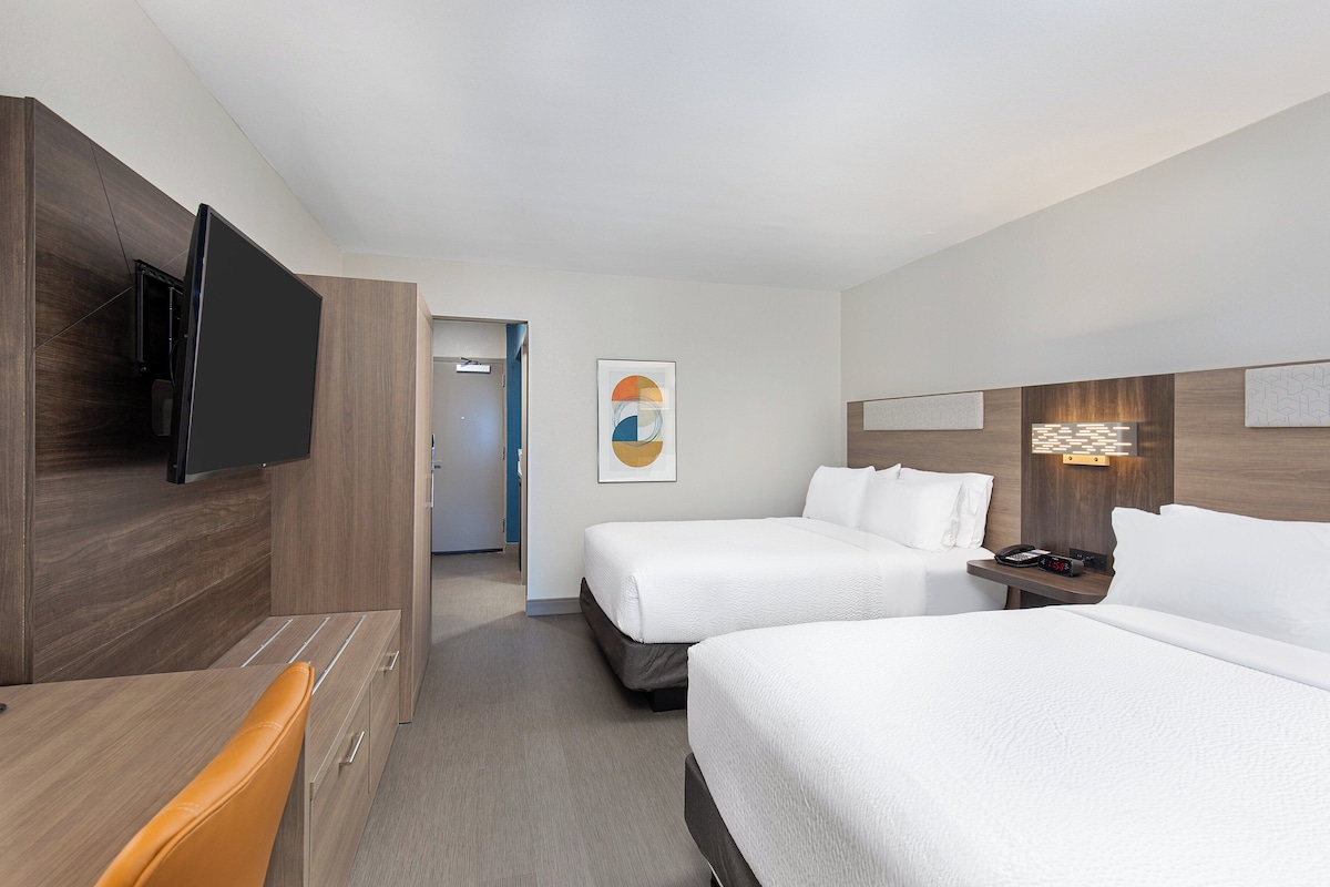 A hotel room with two beds, a TV mounted on the wall, a desk with a chair, and artwork on the walls.