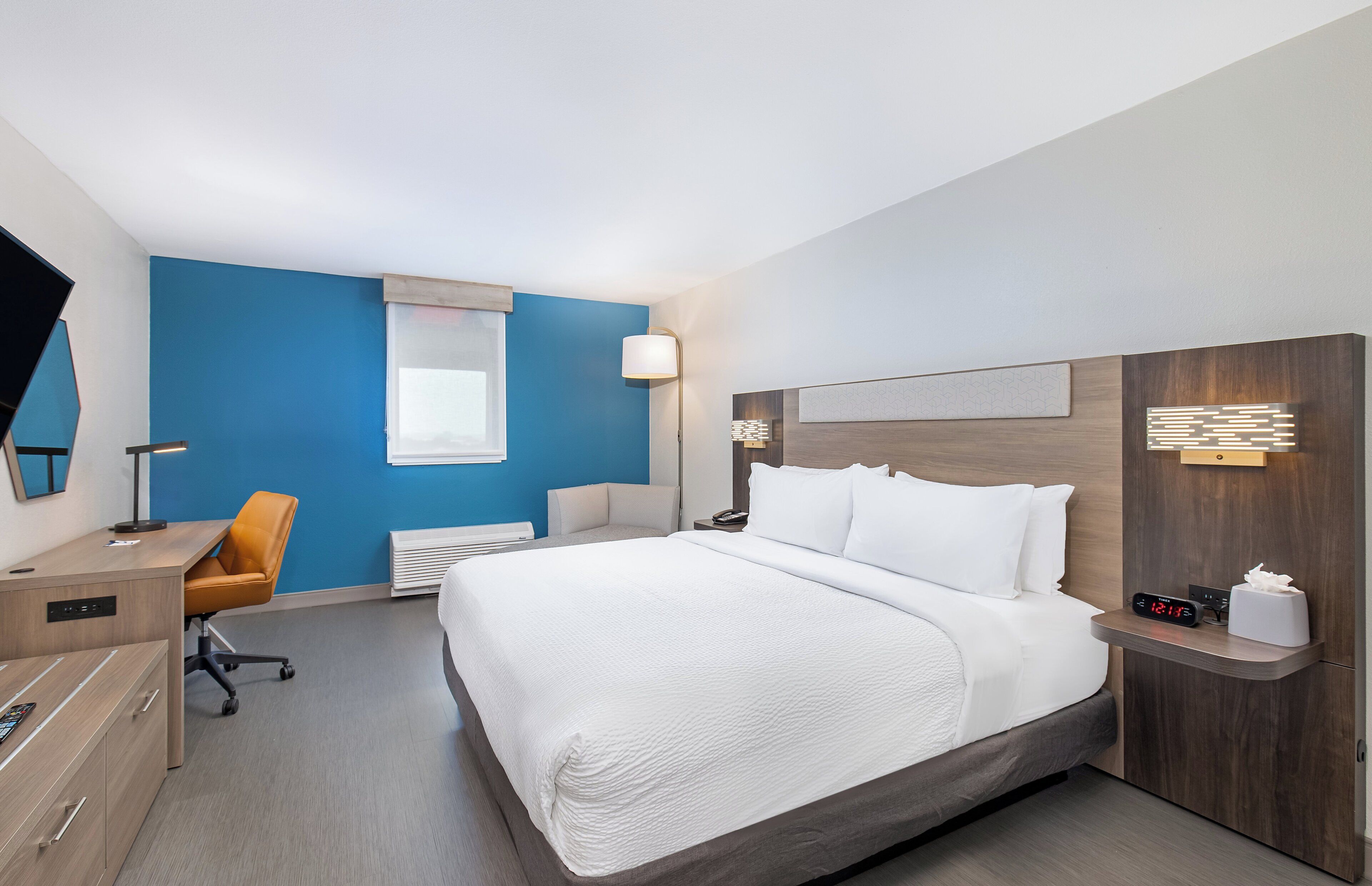 A hotel room with a bed, a desk, a chair, a bedside table, a nightstand, a lamp, a clock, and a window.