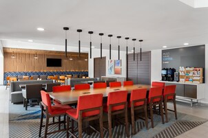 Un comedor de hotel moderno con una mesa larga y sillas rojas, una zona de cocina con estación de café, y un área de descanso con sofás y mesas.