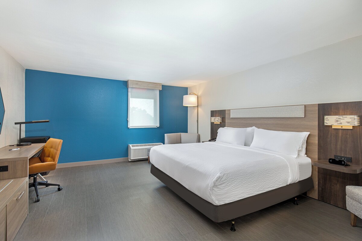 A hotel room with a large bed, a desk, an office chair, a desk lamp, and a bedside table.