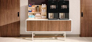 A coffee station with two coffee machines, a shelf with coffee cups and a kettle, and a storage area with boxes and containers. - Holiday Inn Express Atlanta Airport - North by IHG (College Park)