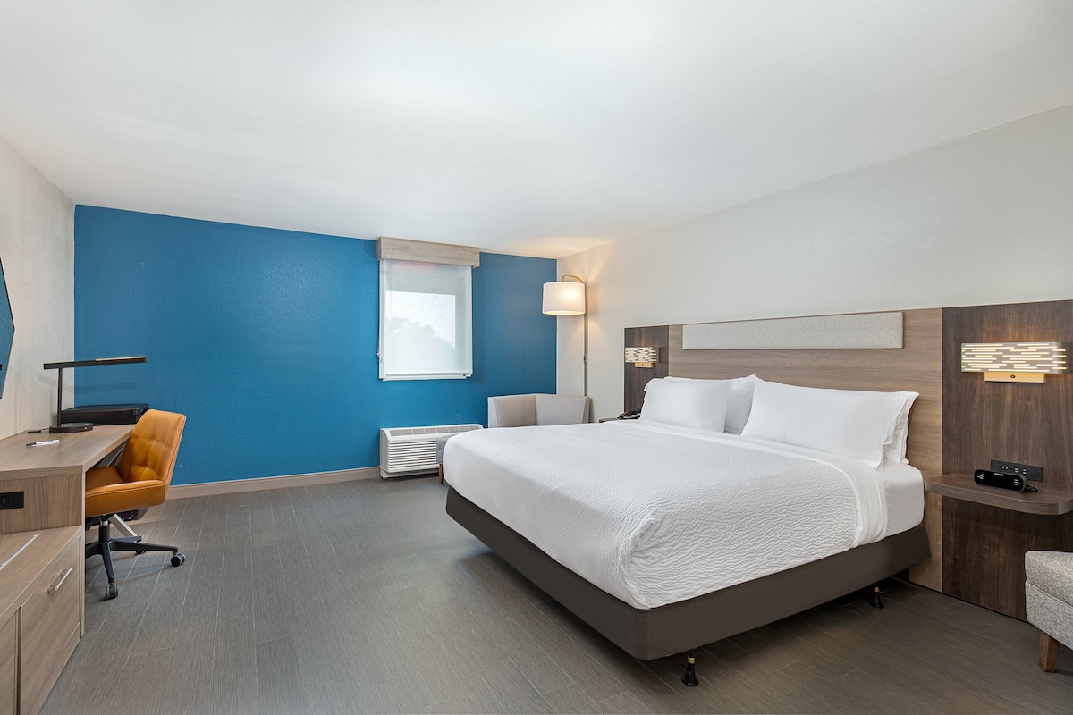 A hotel room with a large bed, a desk, an office chair, a desk lamp, and a bedside table.