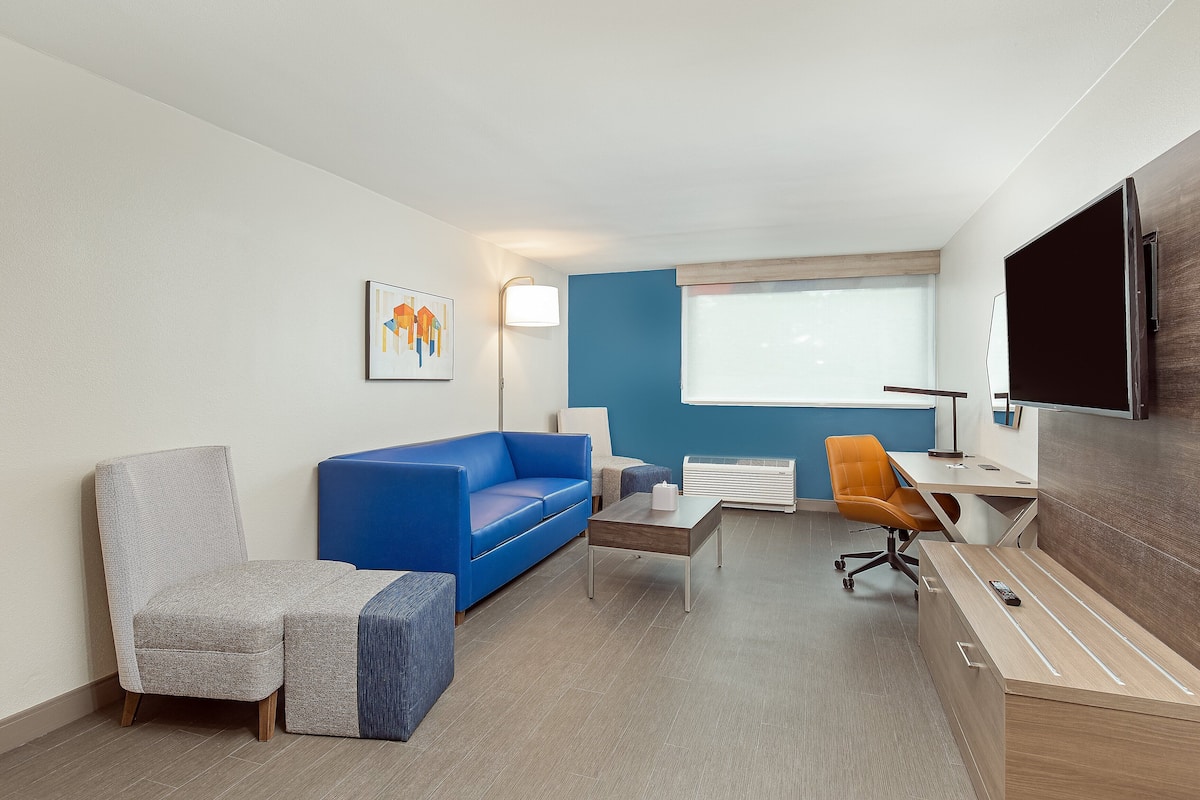 A modern hotel room with a blue sofa, a grey armchair, a wooden desk, and a flat-screen TV mounted on the wall.