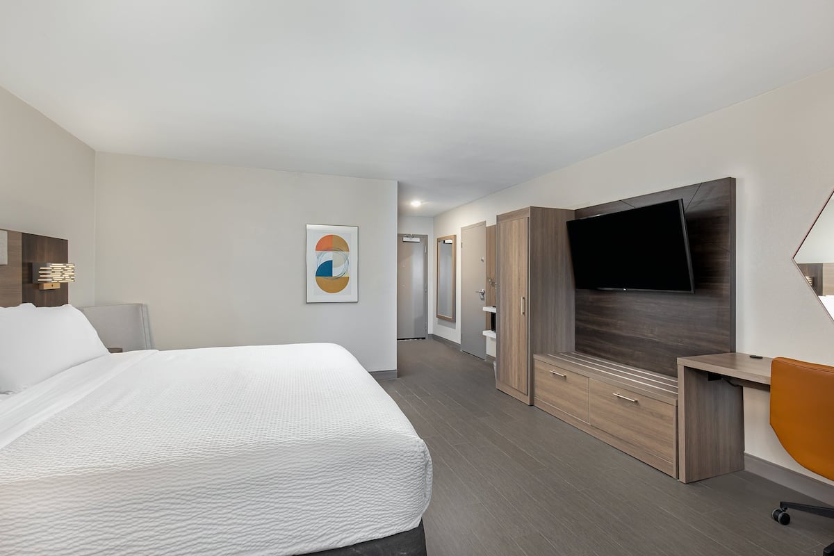 A hotel room with a large bed, a flat-screen TV mounted on the wall, a wooden wardrobe, and a desk with a chair.