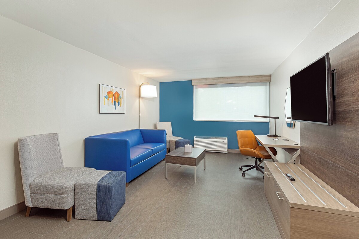 A modern hotel room with a blue sofa, a grey armchair, a wooden desk, and a flat-screen TV mounted on the wall.