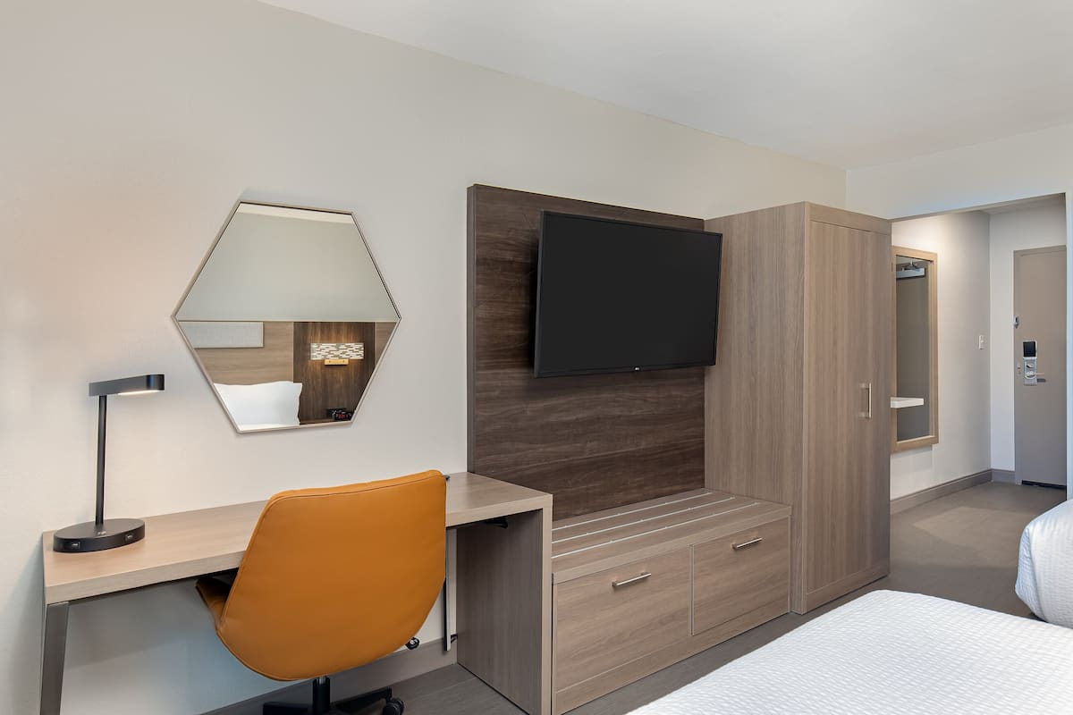 A modern hotel room with a desk, chair, bed, wardrobe, and a flat-screen TV mounted on the wall.