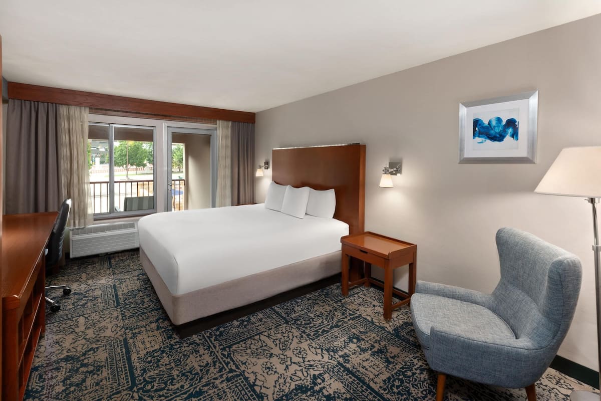 A hotel room with a large bed, a desk, a chair, a window with curtains, a wall-mounted lamp, and a framed picture.