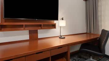 A hotel room with a wooden desk, a television mounted above it, a desk lamp, a chair, and a patterned carpet.