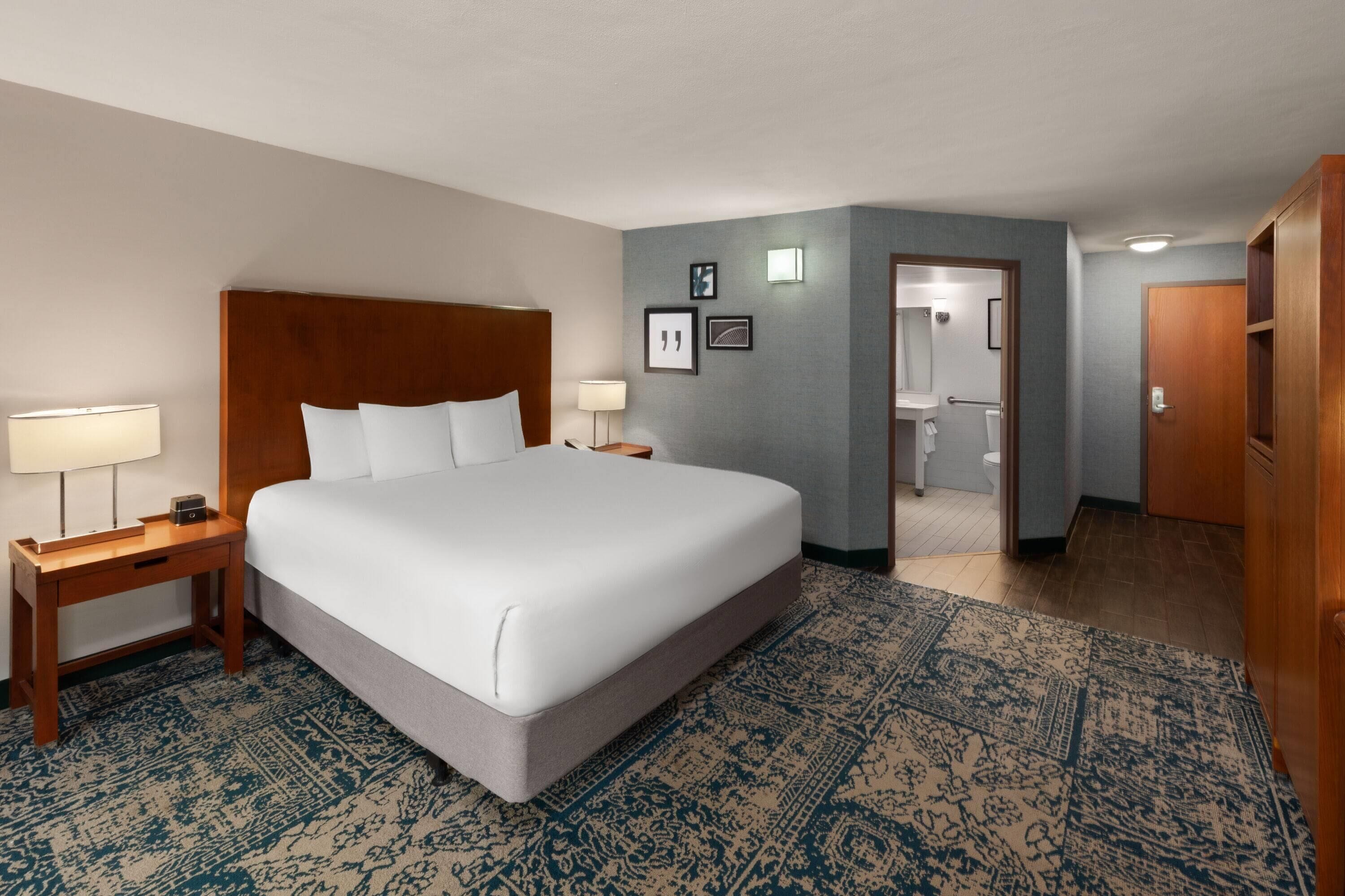 Une chambre d’hôtel avec un grand lit, une table de chevet avec une lampe, un tapis à motifs et une salle de bain visible à travers une porte ouverte.