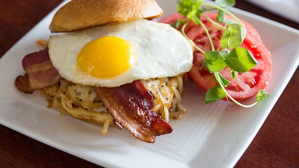 A breakfast sandwich with bacon, egg, and hash browns on a white plate. - The Shores Resort & Spa (Daytona Beach Shores)