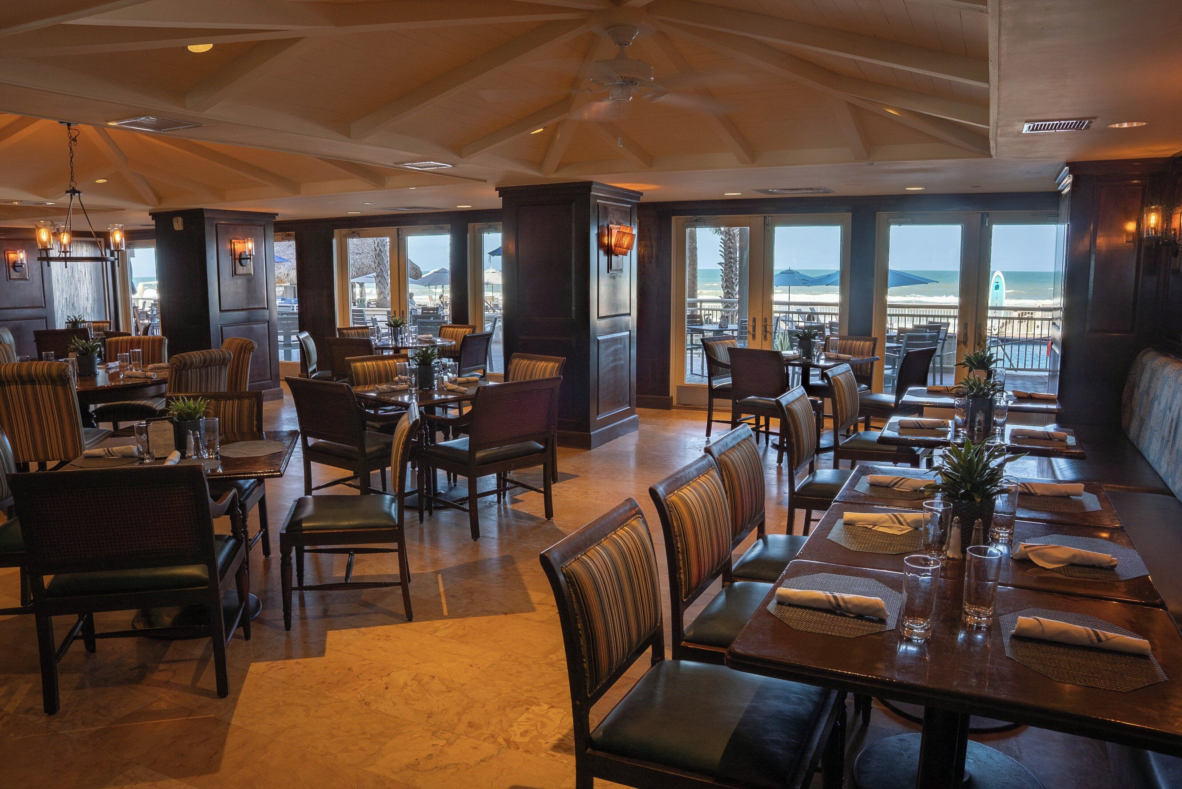 Un restaurant doté de plusieurs espaces de restauration, de grandes fenêtres offrant une vue sur l’océan et d’un ventilateur de plafond.