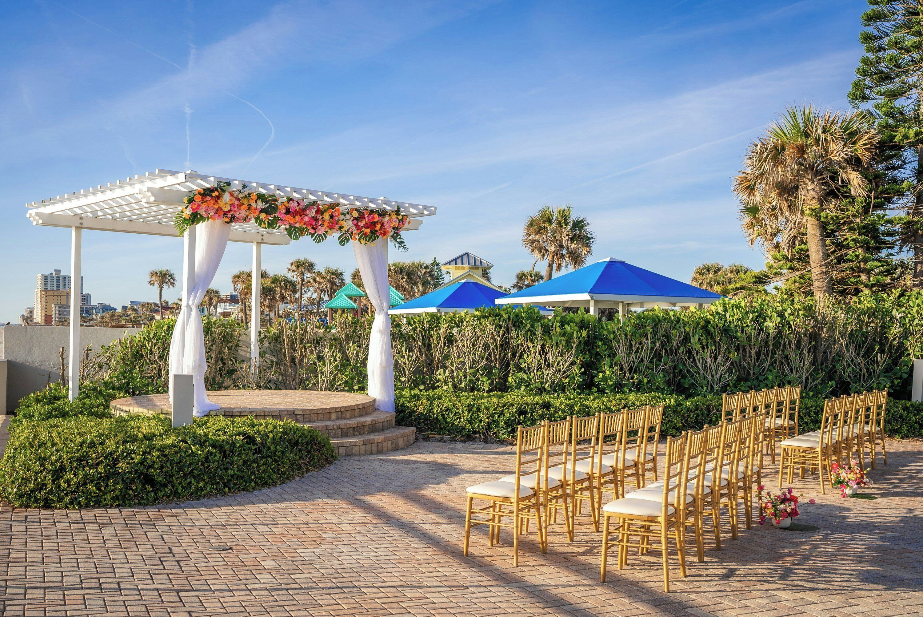 Montaje para una boda al aire libre con un arco de madera blanca, adornos florales y filas de sillas de bambú.