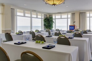 A conference room with tables set for a meeting, large windows with a view of the sea, and a podium with a microphone.