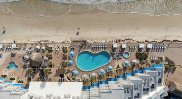 Vista aérea de un complejo turístico con piscina en forma de corazón, tumbonas, sombrillas y una zona de playa.