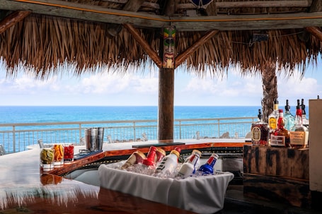 A bar area with a view of the ocean, a thatched roof, and a wooden bar counter with bottles.
