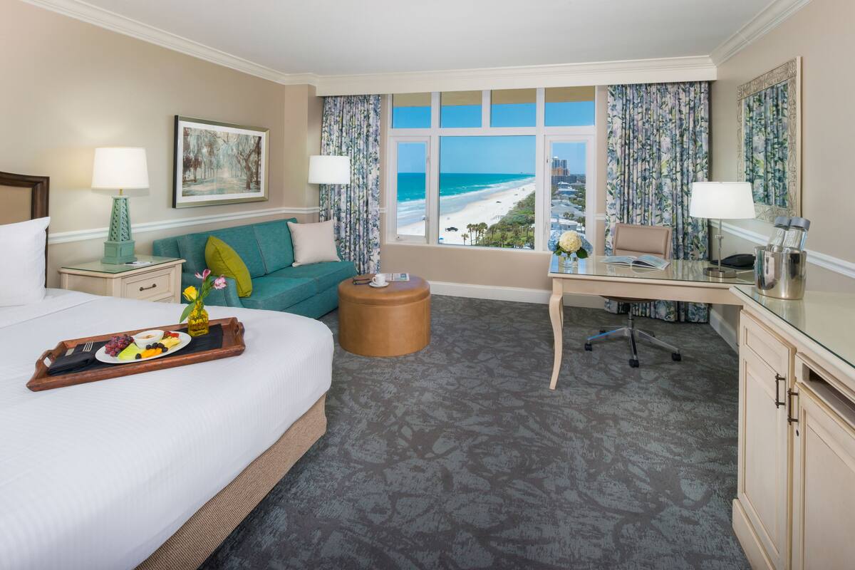 A hotel room with a bed, a desk, a view of the beach, and a dining area with a tray of food.