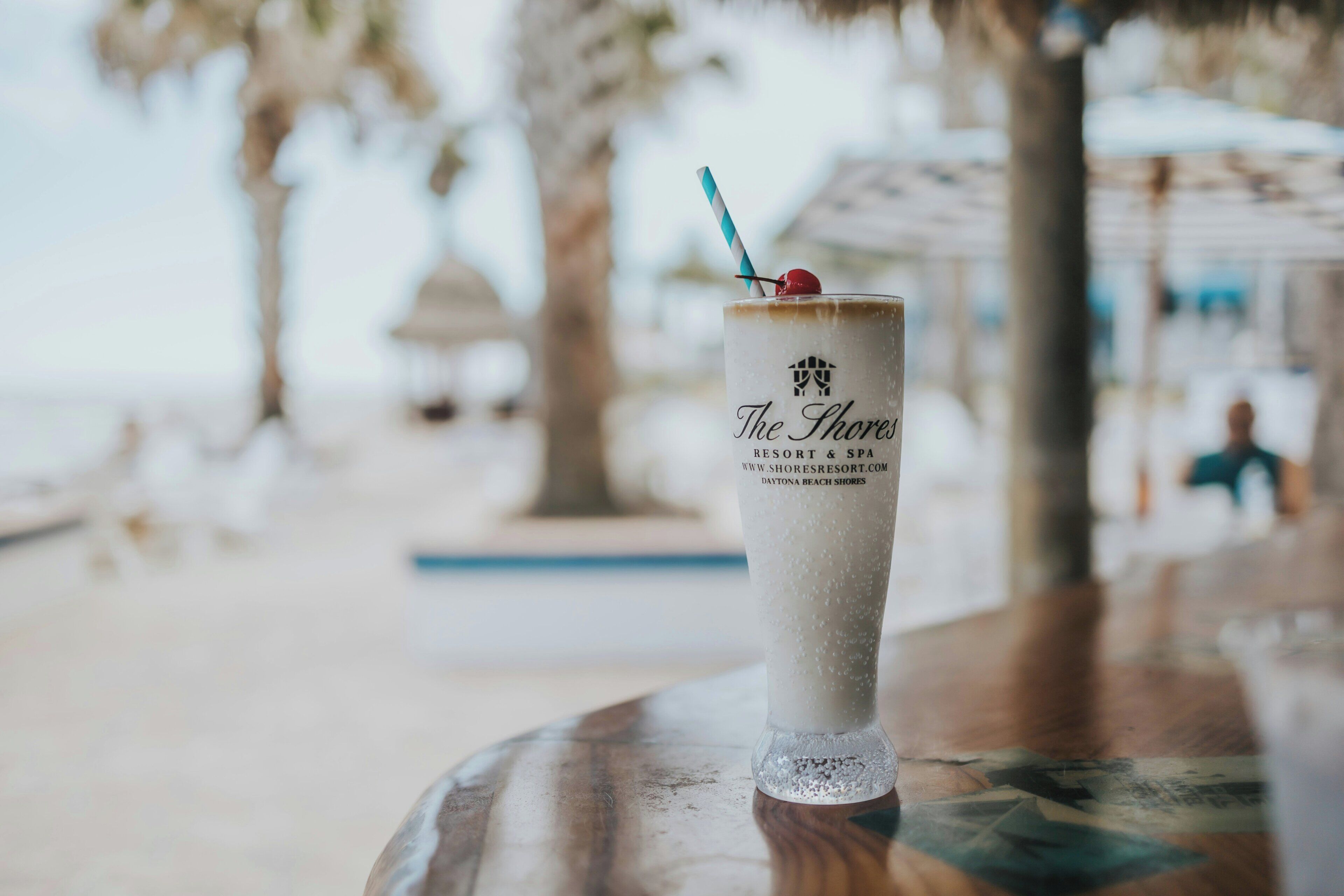 A glass of a beverage with a straw and a cherry on a table at a beachside location.